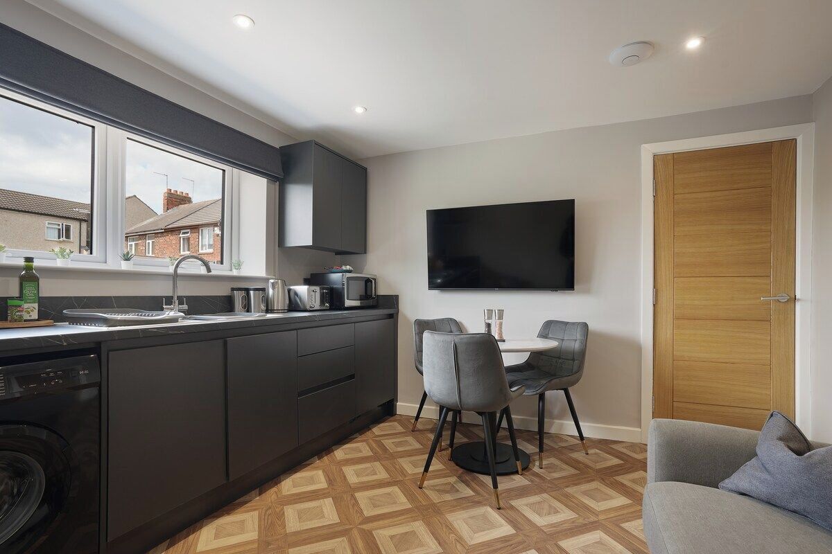 A compact kitchen with modern appliances, a dining area with chairs, a flat-screen TV mounted on the wall, and a wooden door.