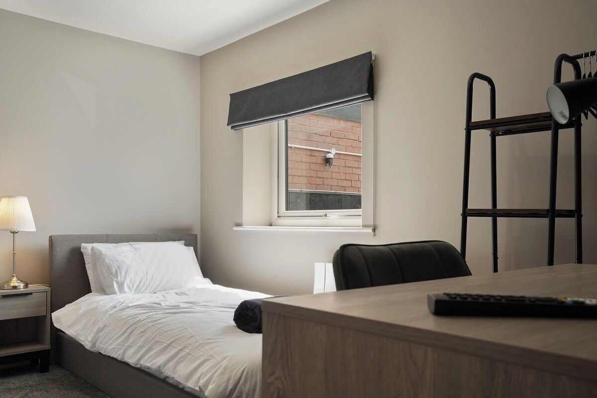 A hotel room with a bed, a desk, a chair, a lamp, and a window with a view of a brick building.