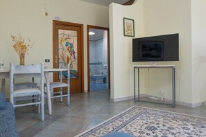 A living room with a TV on a stand, a dining area with chairs, a rug, and a door leading to a bathroom.