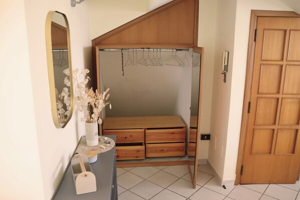 A bedroom with a wooden wardrobe, a mirror, a vase with dried flowers, and a small table with drawers.