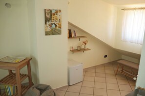 Een knus leefruimte met een kleine koelkast, een houten tafel met een tekening, een bankje en een schilderij van een brug aan de muur.