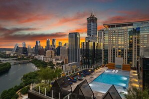 Ein Stadt-Skyline bei Sonnenuntergang mit einem Poolbereich, Liegestühlen und Blick auf den Fluss.