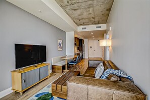 A modern living room with a flat-screen TV, a sofa, a dining area, and a kitchen in the background. - 24th-floor 1BR Sleeps 4 | Rainey + Rooftop Pool (Austin)