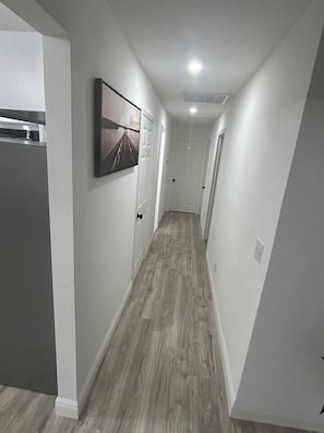 A narrow hallway with a wooden floor, white walls, and a framed picture on the wall. - Modern 3BR | Near DT & Galleria (Houston)