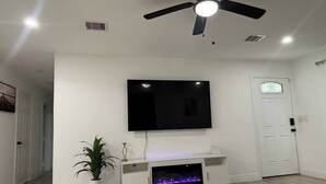 Un salon doté d’un téléviseur à écran plat fixé au mur, d’un ventilateur de plafond, d’une cheminée avec un éclairage bleu et d’une porte ornée d’un vitrail décoratif.