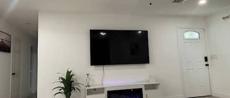 Un salon doté d’un téléviseur à écran plat fixé au mur, d’un ventilateur de plafond, d’une cheminée avec un éclairage bleu et d’une porte ornée d’un vitrail décoratif.