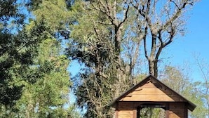 A wooden structure with a sloped roof, possibly a gazebo, surrounded by grass and trees.