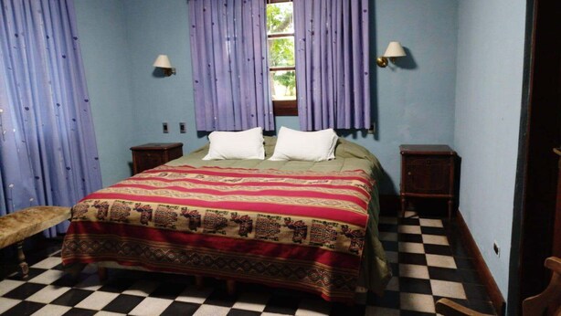 A room with a checkered floor, a bed with patterned bedding, two nightstands, a bench, and purple curtains.