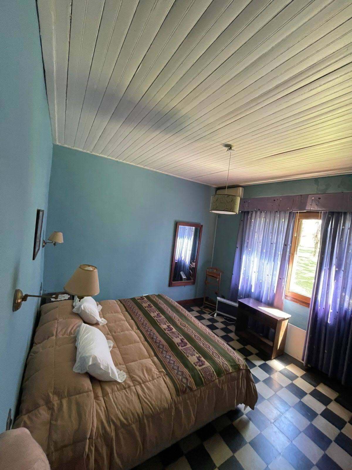 Una habitación con cama, escritorio, silla, lámpara, ventana con cortinas y piso a cuadros.