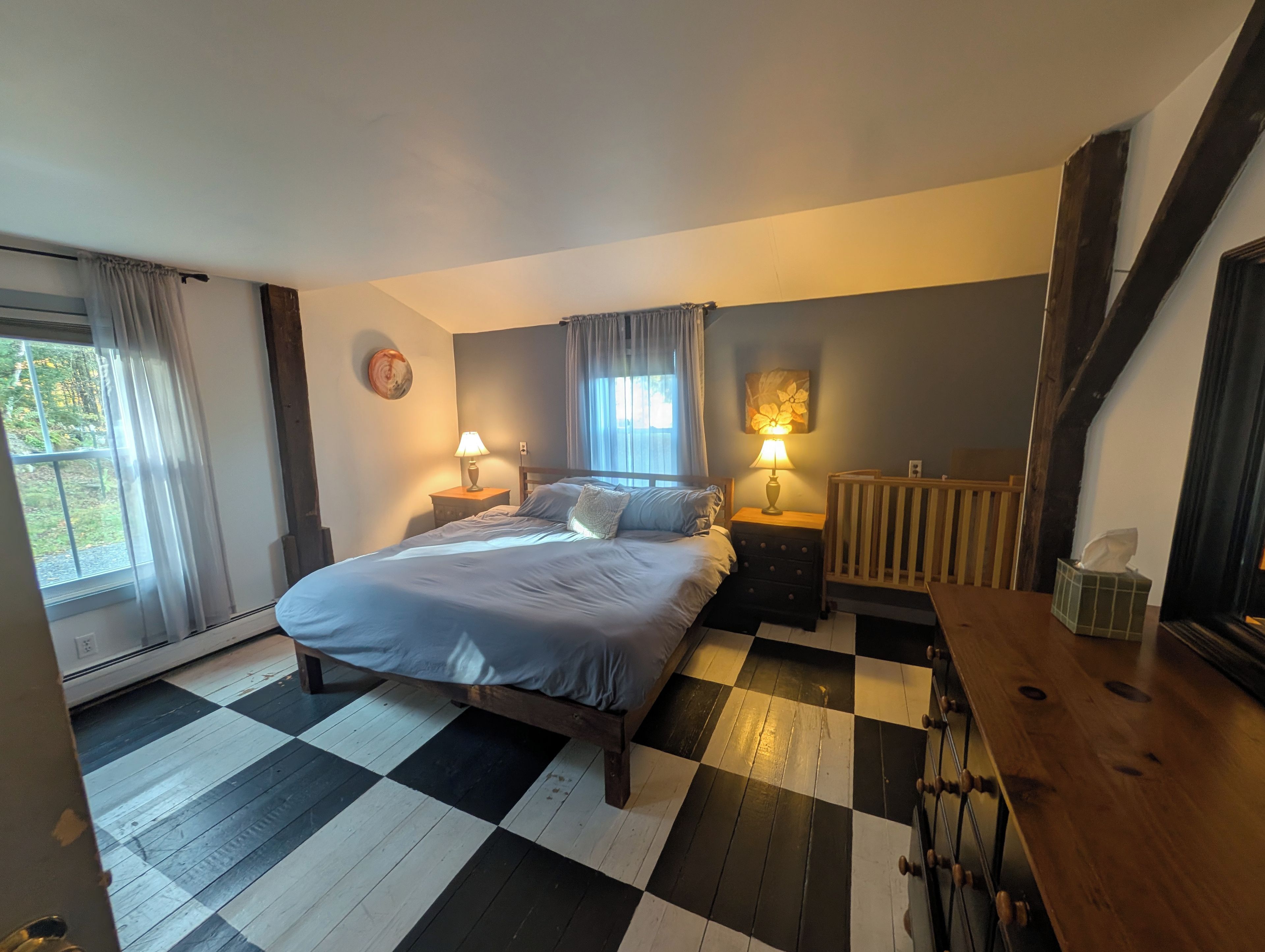 A bedroom with a checkered floor, a bed with pillows, two bedside tables with lamps, a crib, and a radiator.