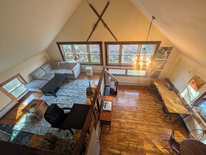 A loft-style room with a high ceiling, wooden flooring, a sofa, a dining table with chairs, and large windows offering a view of trees.