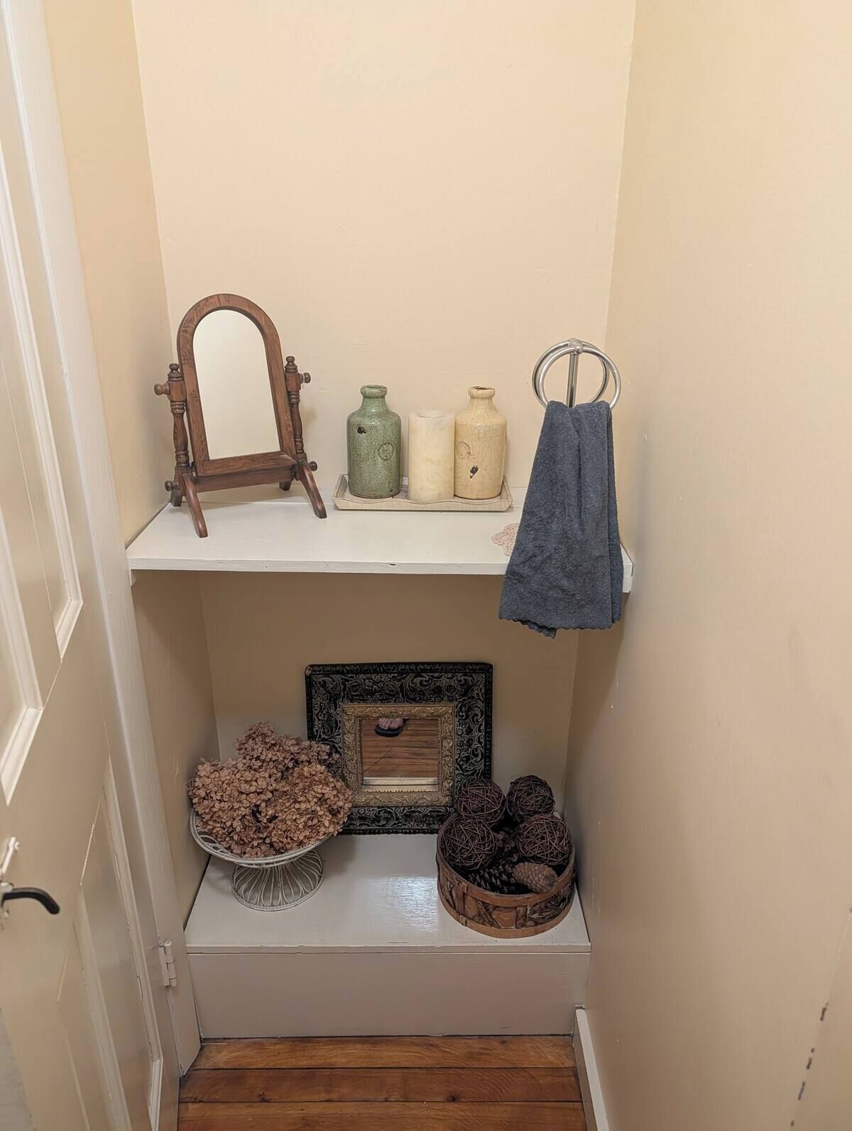 Un couloir étroit où se trouve un meuble en bois sur lequel sont posés un miroir et trois pots en céramique, placés sur une étagère. En dessous, un meuble blanc expose des fleurs séchées et des boules décoratives.