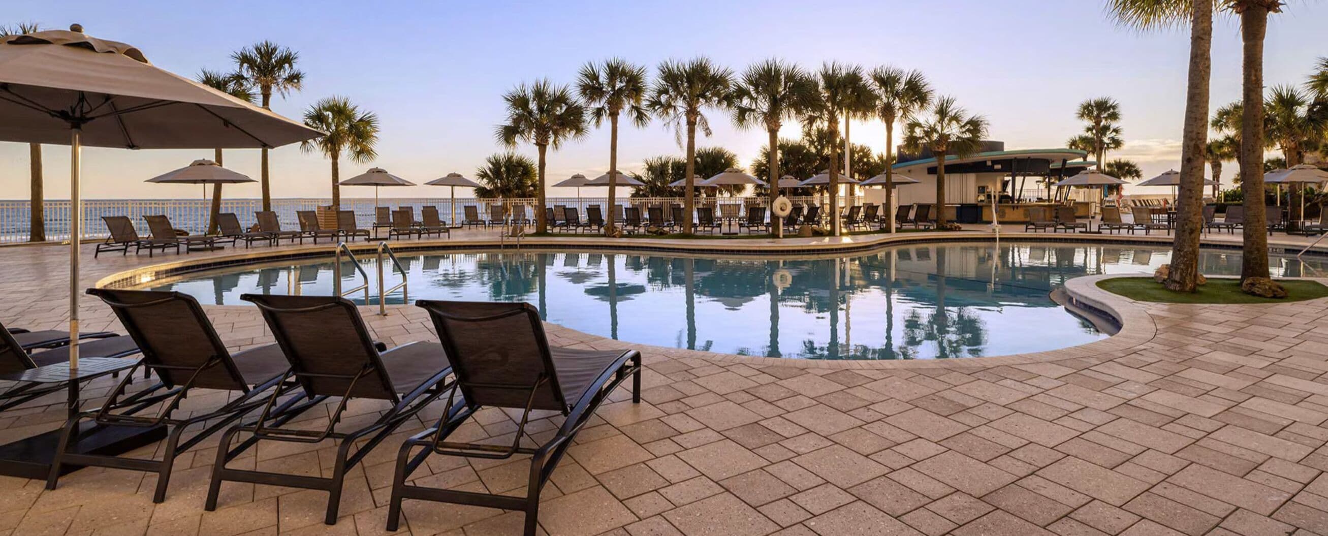 A poolside area with lounge chairs and umbrellas, surrounded by palm trees.