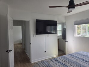 Une chambre à coucher avec un lit, un ventilateur de plafond, un téléviseur fixé au mur et une fenêtre avec des stores.