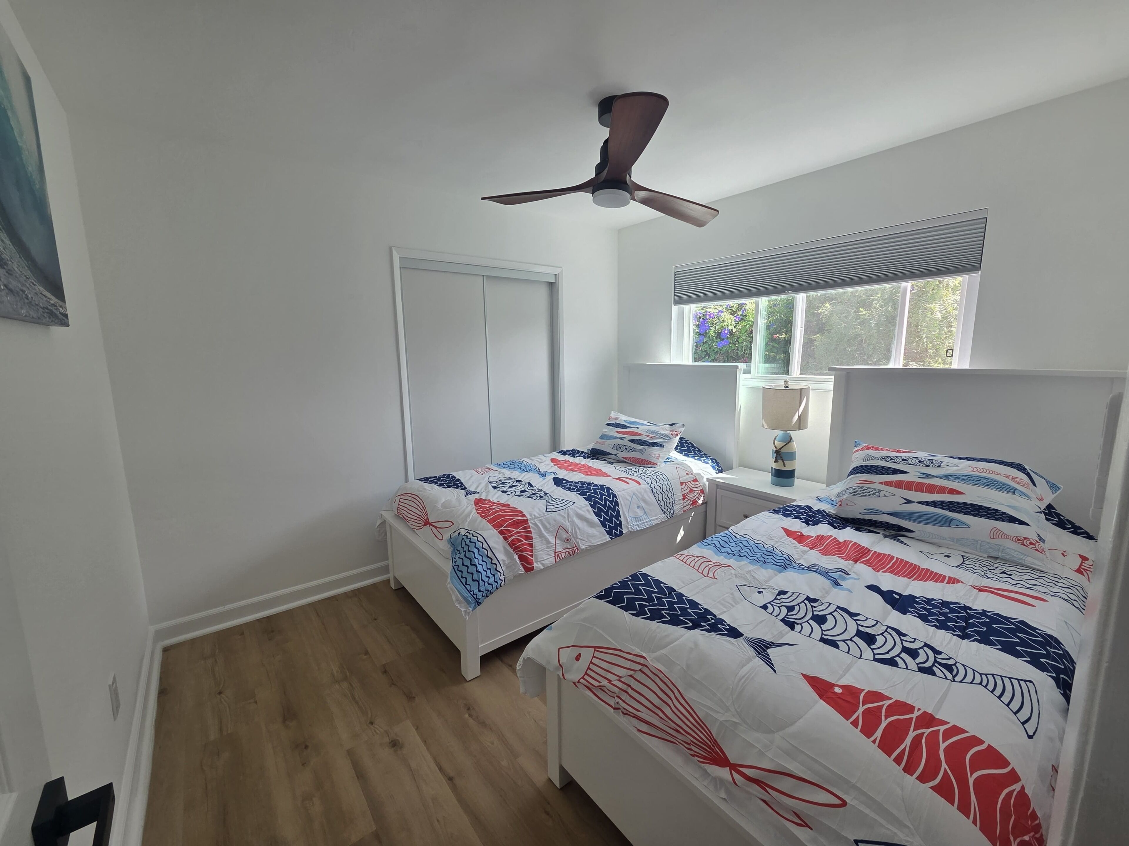 A bedroom with two single beds, a ceiling fan, a window with blinds, and a door.