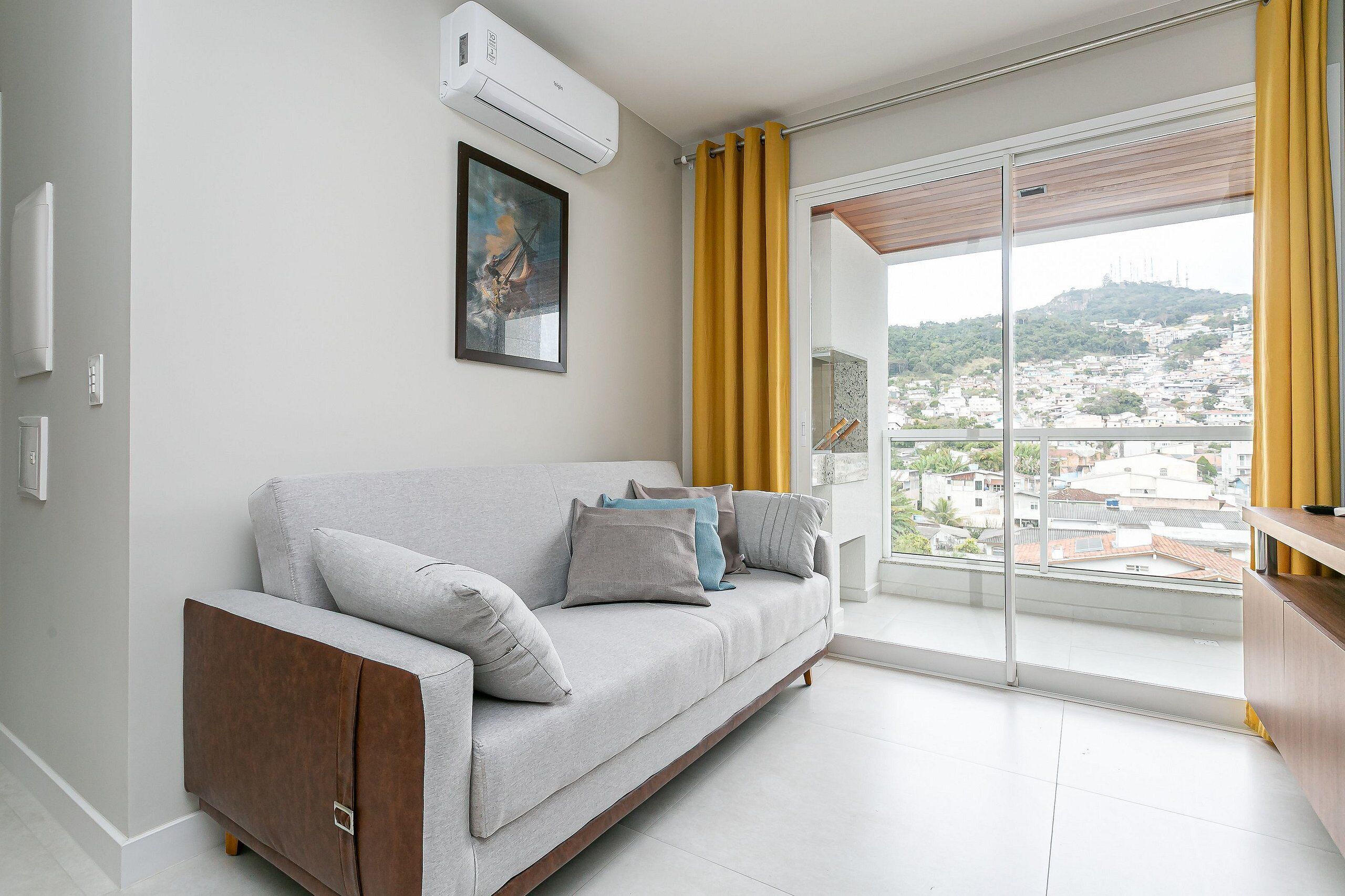 Uma sala de estar moderna com um sofá cinza, um ar condicionado fixo, uma obra de arte emoldurada e vista para um horizonte urbano através de janelas amplas com cortinas amarelas.
