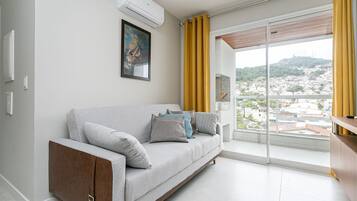 Uma sala de estar moderna com um sofá cinza, um ar condicionado fixo, uma obra de arte emoldurada e vista para um horizonte urbano através de janelas amplas com cortinas amarelas.