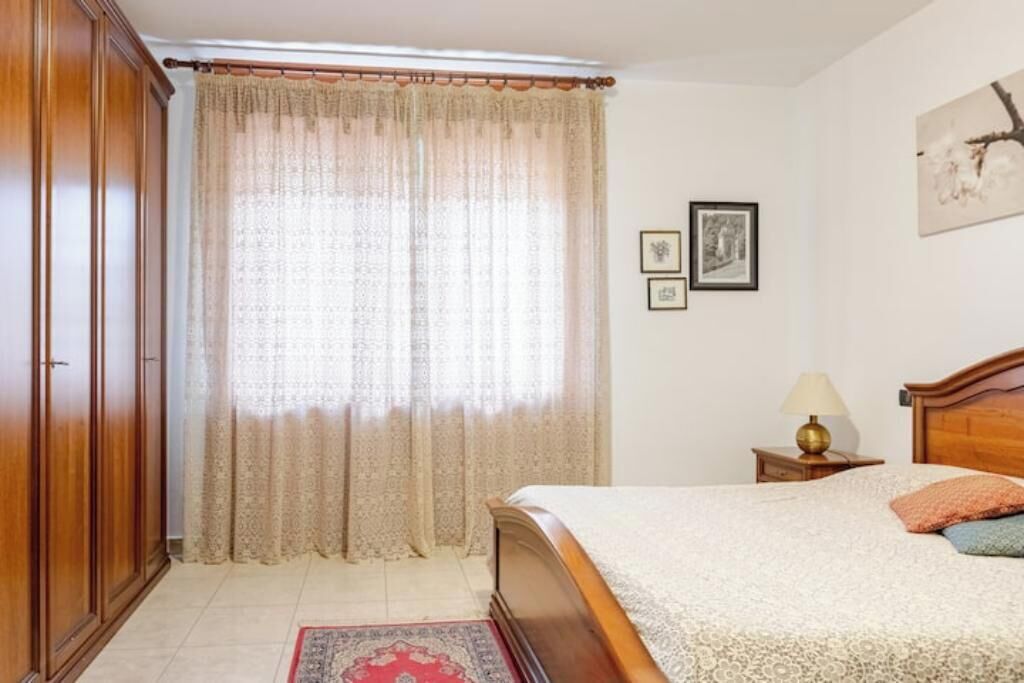 A bedroom with a bed, a nightstand, a lamp, a wardrobe, and a window with curtains.