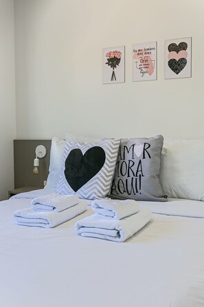 Een net opgemaakt bed met opgevouwen witte handdoeken, een kussen met een hartdessin en drie wanddecoraties met tekst en hartmotieven.