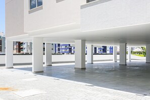 A spacious covered outdoor area with a tiled floor, supported by white pillars, and a view of a modern building with blue accents in the background. - 500m from the sea | pool | Barbecue grill #IA38 (Florianópolis)