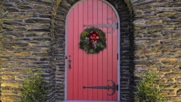 A stone building with a red door, a wreath, and a metal handle.