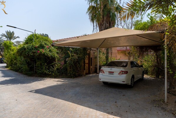 A white car parked under a covered area with a tiled roof, surrounded by greenery and a clear sky.