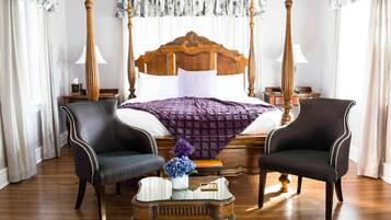 A four-poster bed with a wooden headboard, two upholstered armchairs, a small side table with a vase of flowers, and a patterned rug.