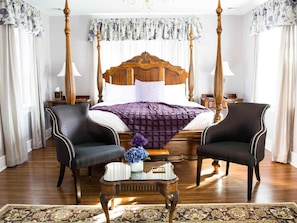 A four-poster bed with a wooden headboard, two upholstered armchairs, a small side table with a vase of flowers, and a patterned rug.