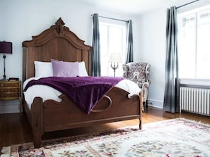 A bedroom with a wooden bed, a purple throw blanket, a floral-patterned rug, a nightstand with a lamp, and a chair. - The Wilson Suite at The Manor on Front (Harrisburg)