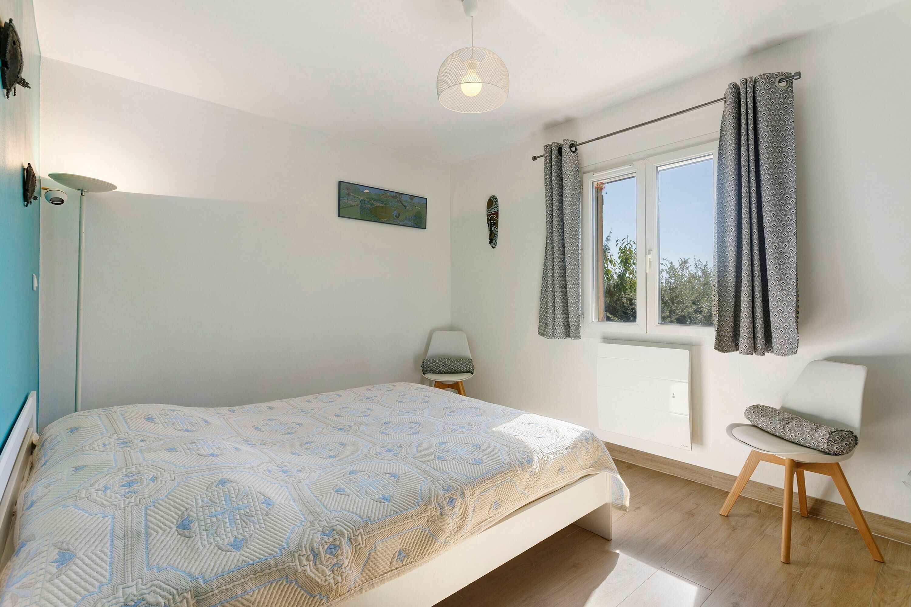 A bedroom with a bed, a window with curtains, a chair, a lamp, and a painting on the wall.