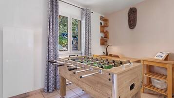 Et værelse med et bordtennisbord, et træskrivebord, en stol, et vindue med gardiner og en reol monteret på væggen.