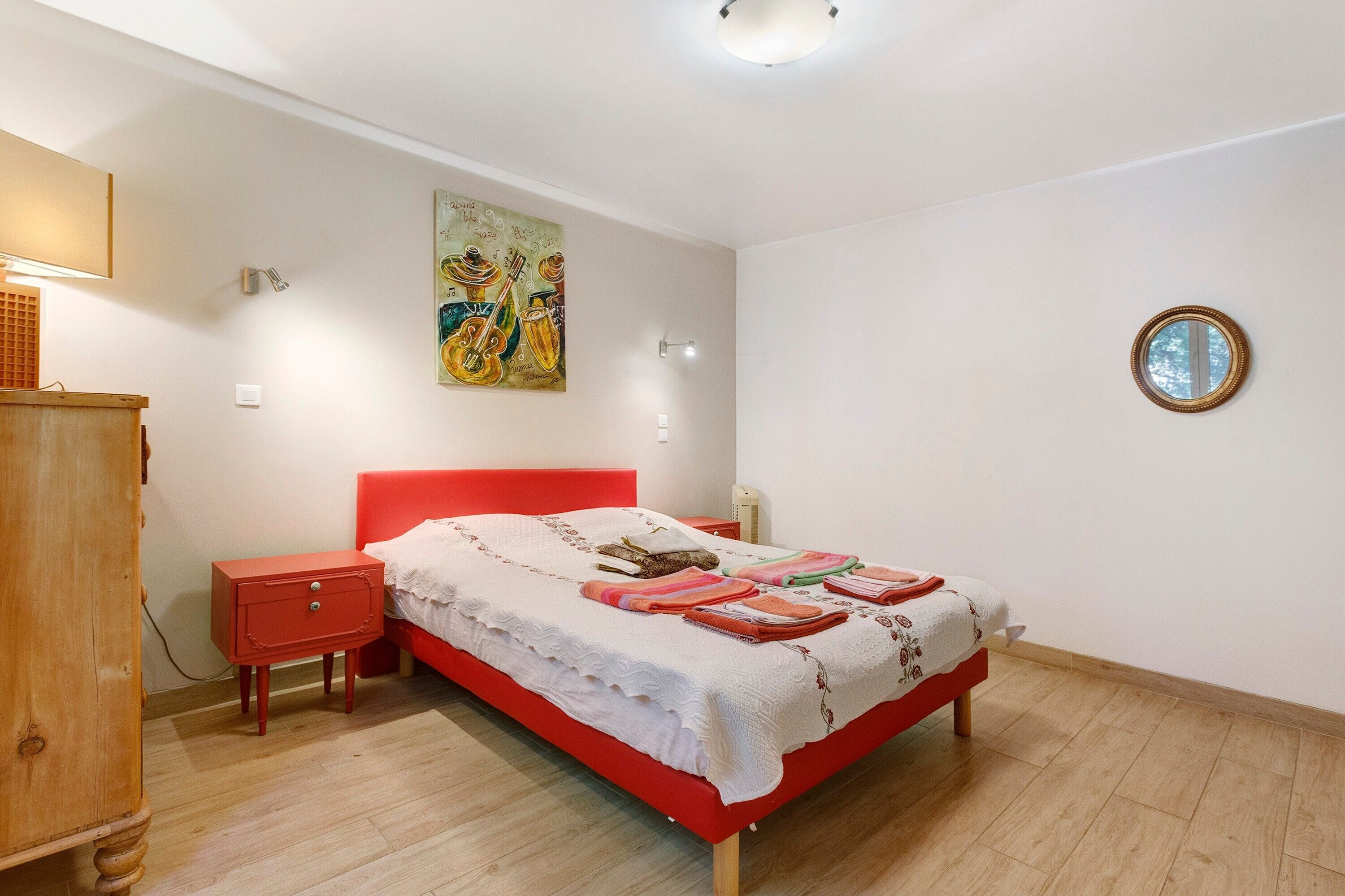 Une chambre à coucher avec un lit rouge, une table de chevet en bois, une commode, une lampe fixée au mur et un tableau.