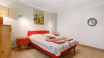 Une chambre à coucher avec un lit rouge, une table de chevet en bois, une commode, une lampe fixée au mur et un tableau.