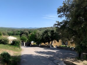 Une allée pavée mène à un bâtiment à la façade beige, entouré d’arbres et situé dans un paysage vallonné.