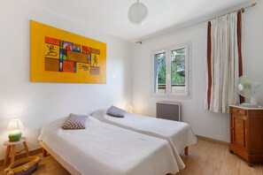Une chambre avec deux lits, une fenêtre avec des rideaux, un radiateur, une lampe, un ventilateur et un tableau au mur.