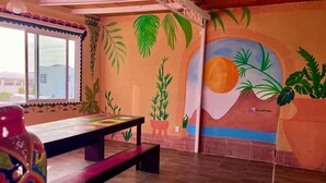 Um quarto com piso de madeira, uma mesa com tampo colorido, um banco e um mural com plantas tropicais e um sol.