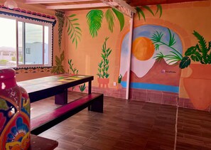 Ein Zimmer mit Holzboden, einem Tisch mit bunter Tischplatte, einer Bank und einem Wandbild mit tropischen Pflanzen und einer Sonne.