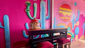 Quarto com uma parede rosa vibrante com um mural de cactos, uma mesa de madeira com almofadas vermelhas e um banco de madeira.