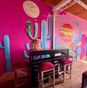 Ein Zimmer mit einer leuchtend rosa Wand, die ein Kaktus-Wandbild zeigt, einem Holztisch mit roten Kissen und einer Holzbank.