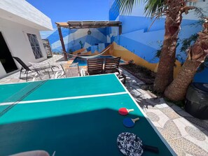 Ein Billardtisch mit zwei Spielschlägern und einem Tischtennis-Schläger, umgeben von Stühlen und einer Palme.