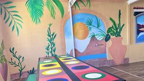 Um mural colorido com palmeiras e um sol, uma mesa com círculos coloridos e um vaso de planta.