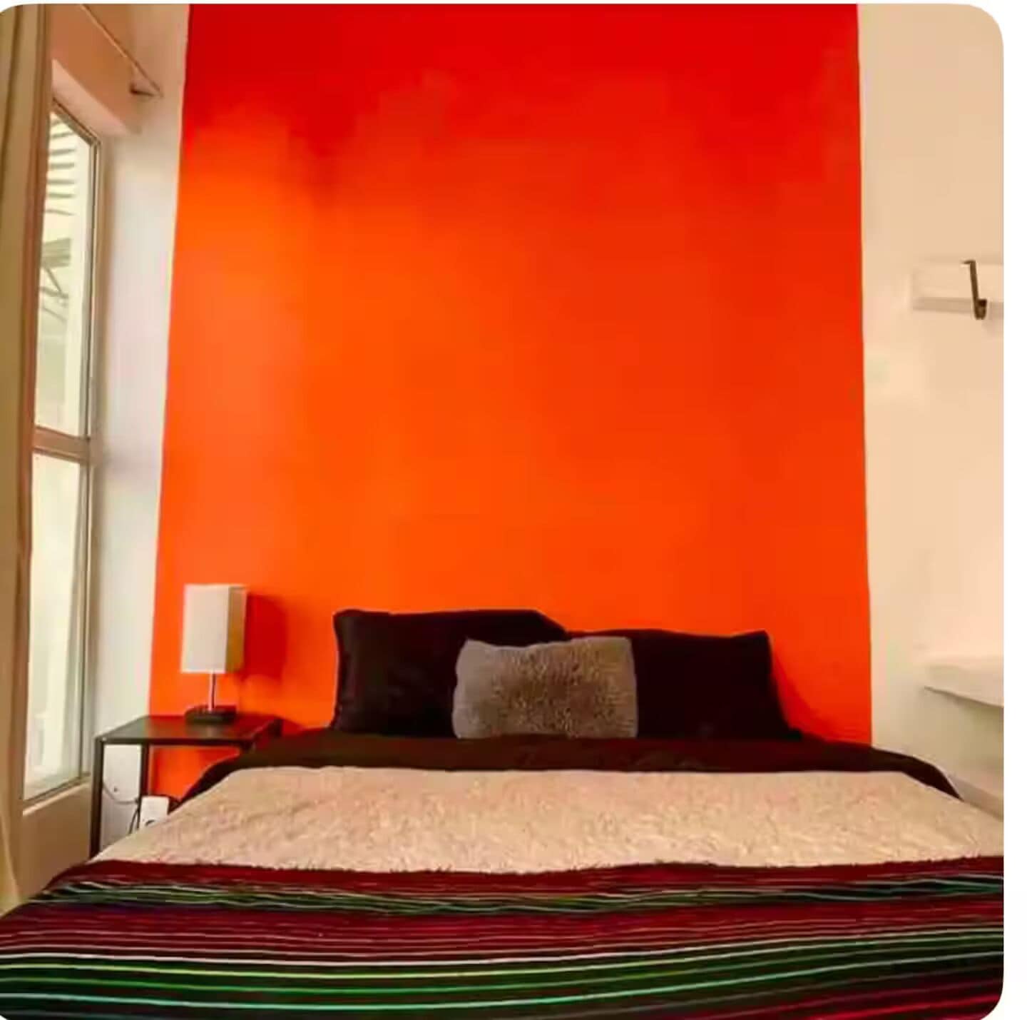 Een kamer met een bed tegen een rode muur, een klein nachtkastje met een lamp, en een raam aan de linkerkant.
