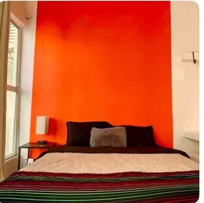 Ein Zimmer mit einem Bett an einer roten Wand, einem kleinen Nachttisch mit einer Lampe und einem Fenster links.