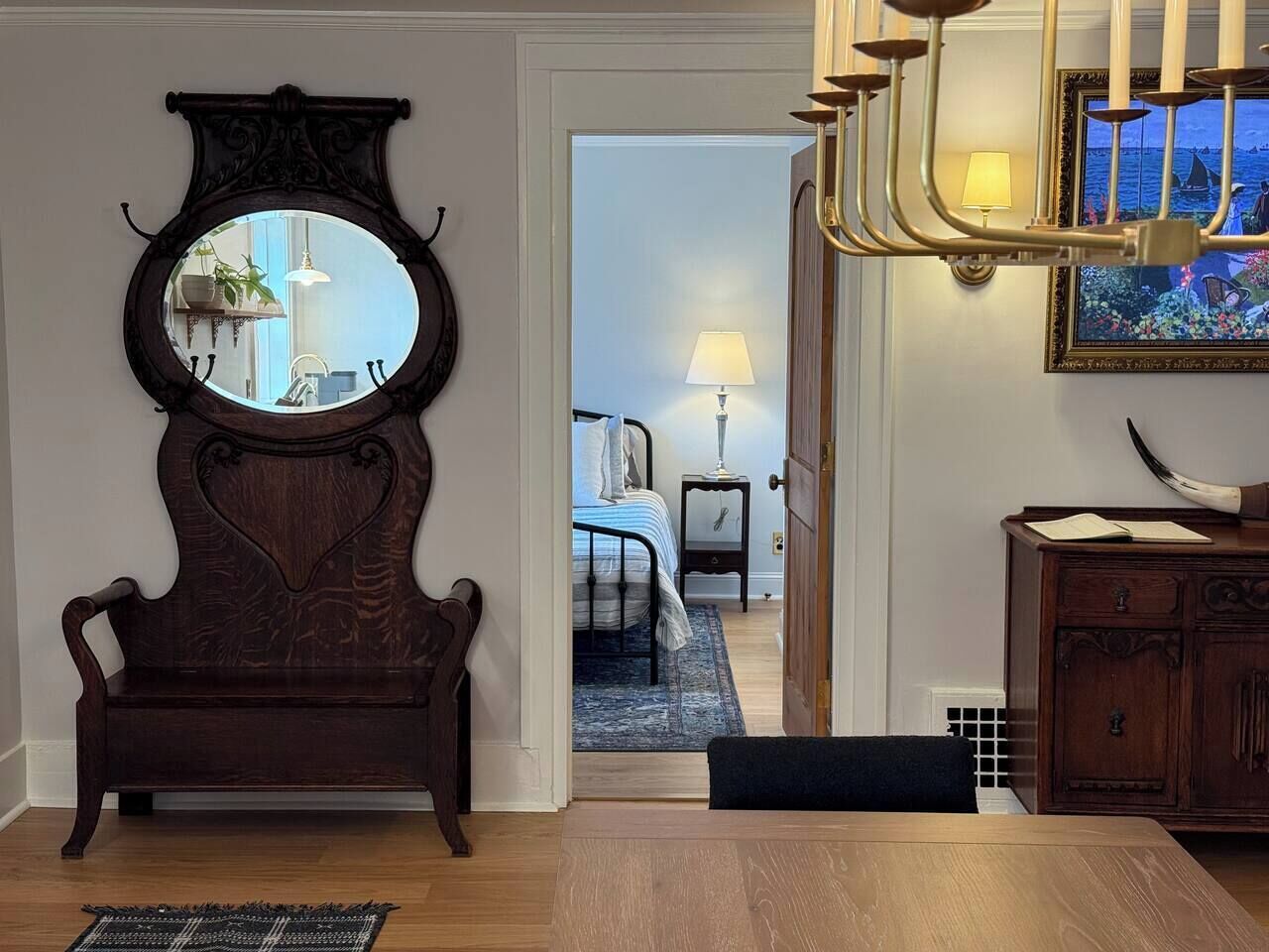 A room with a dark wooden bench, a round mirror, a wooden cabinet, and a doorway leading to a bedroom with a bed and a nightstand.