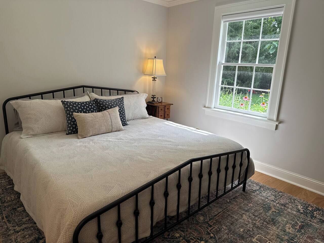 A neatly made bed with a patterned bedspread and pillows, a bedside table with a lamp, and a window with a view of greenery.