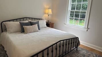 A neatly made bed with a patterned bedspread and pillows, a bedside table with a lamp, and a window with a view of greenery.