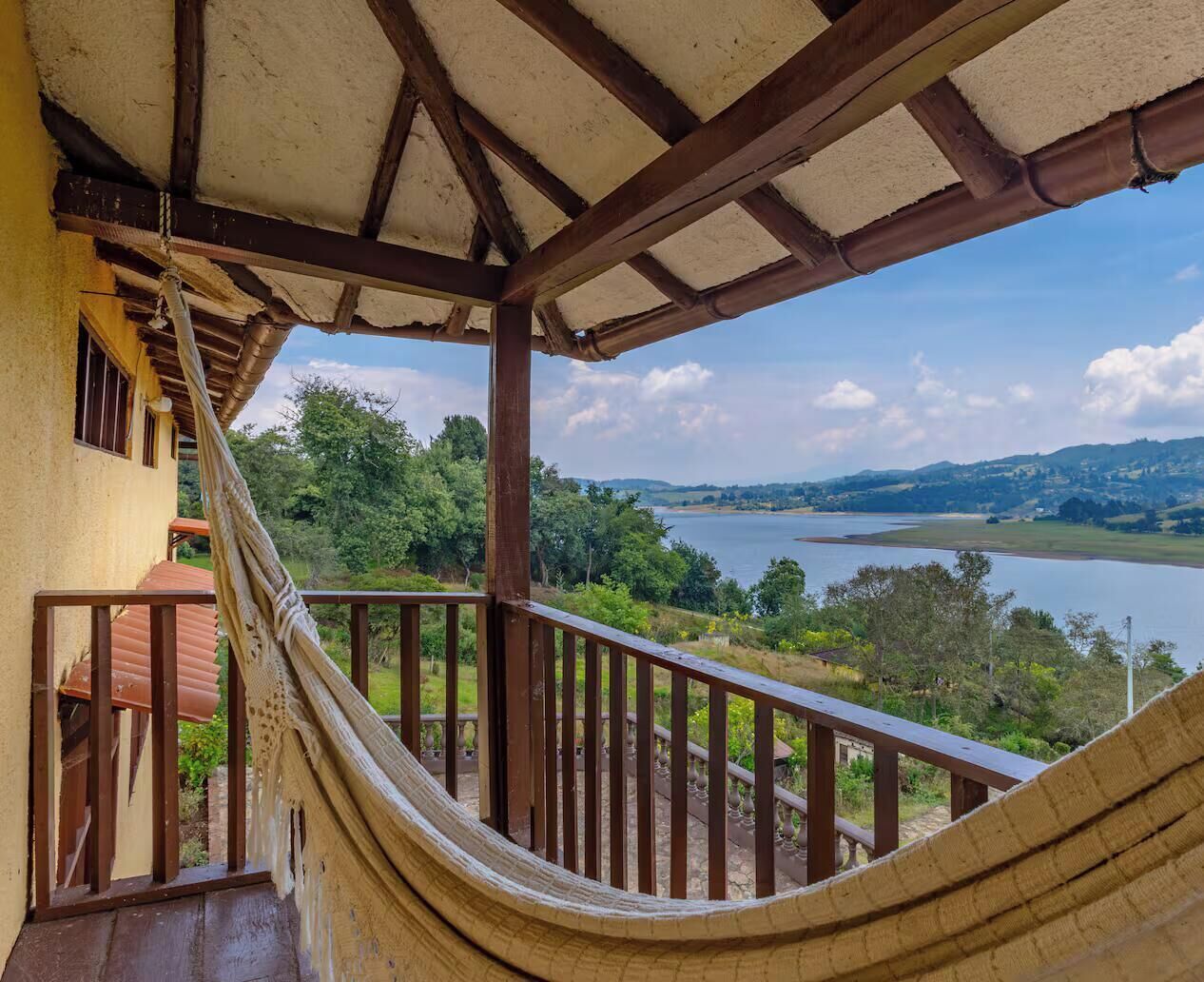 A hammock under a wooden balcony with a view of a lake and hills.