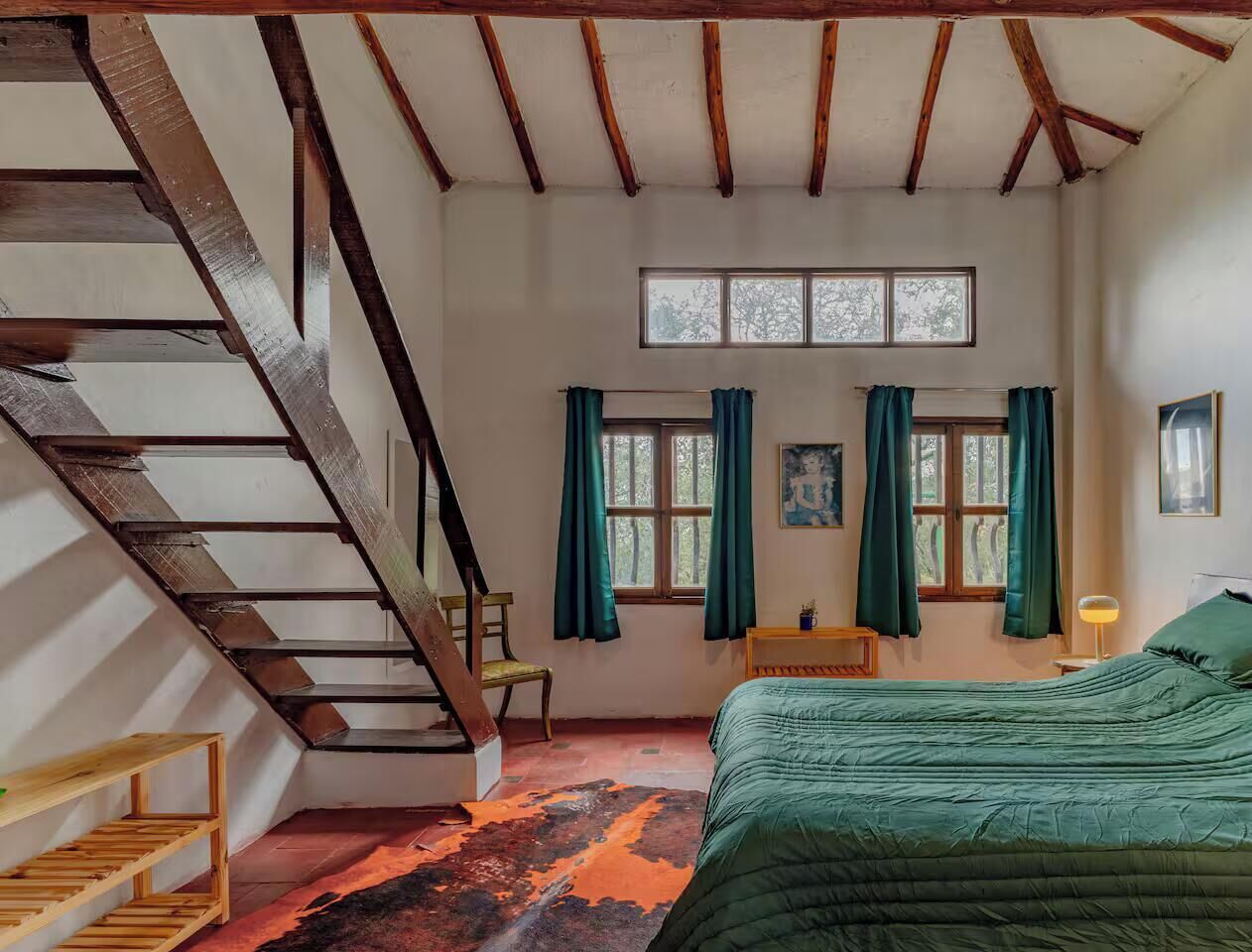 Ein Schlafzimmer mit schrägen Wänden, einem Bett, einem Sessel, einem kleinen Tisch und einem fenster mit Vorhängen.