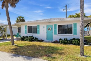 A single-story house with a light-colored exterior, numbered 44, featuring a front porch with a green door and shutters, and a small front yard with grass and shrubs. - Paradise on Pearl 2-bedroom bungalow at Englewood Beach (Englewood)