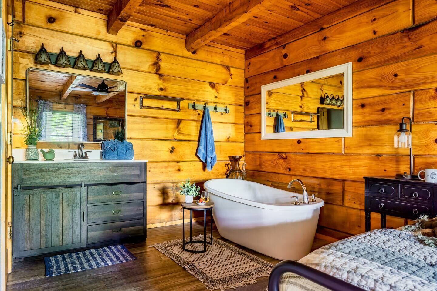 A wooden cabin interior with a bathroom featuring a bathtub, a vanity with a mirror, and a small table with a plant.